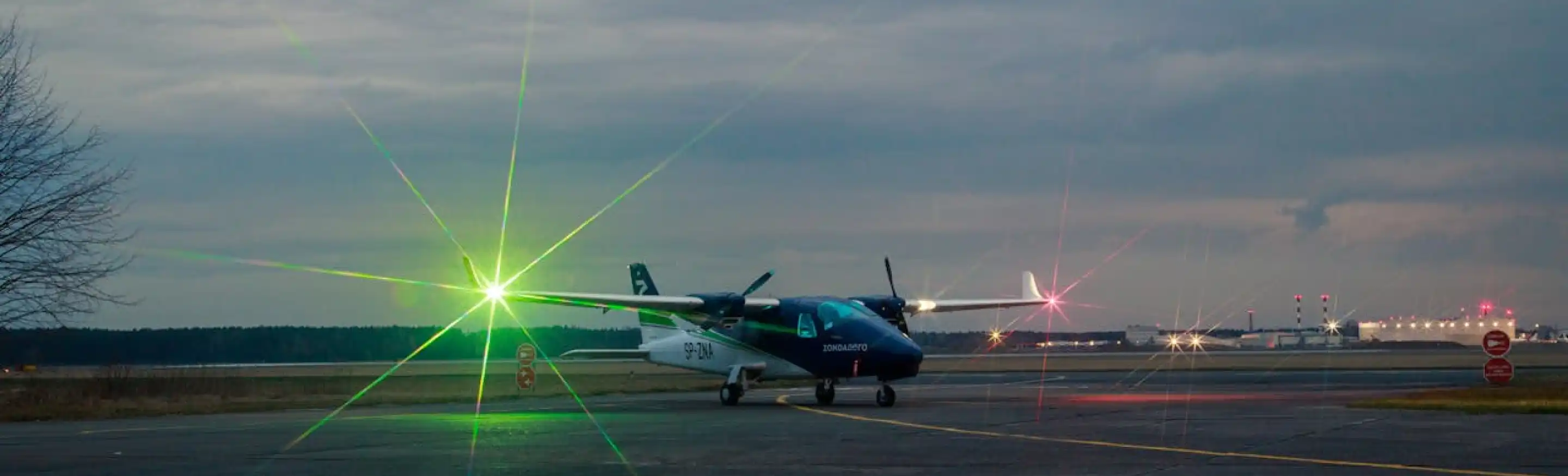 Small propeller airplane with navigation lights on a runway during twilight, with an airport terminal in the background